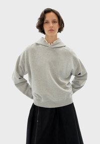Sudadera gris con capucha y un ajuste relajado, con bolsillo frontal tipo pouch y capucha. Presenta una textura suave y puños de canalé. Llevada sobre una falda negra.