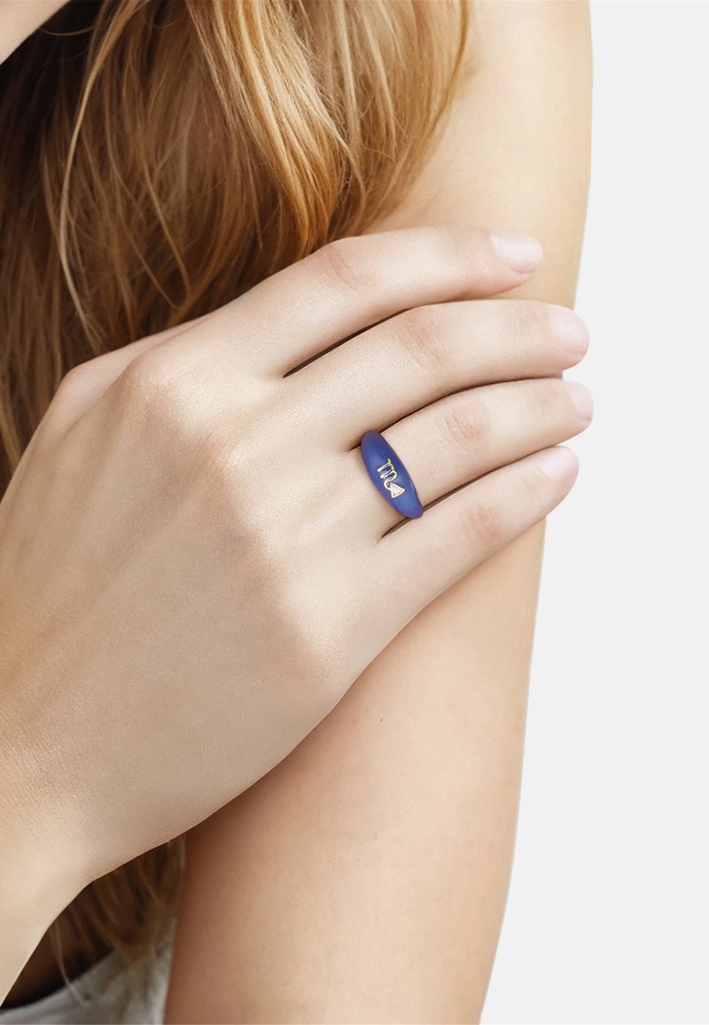 Mano con piel clara que lleva un anillo azul con un símbolo dorado del zodiaco Escorpio, descansando sobre un brazo desnudo con cabello rubio visible.