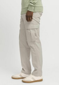 Lyse beige cargobukser med sidelommer, avslappet passform og rette ben, kombinert med hvite joggesko og en grønn genser.