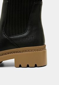 Bottine en cuir noir avec un panneau élastique côtelé, semelle épaisse beige, et détail de logo en relief sur le côté.