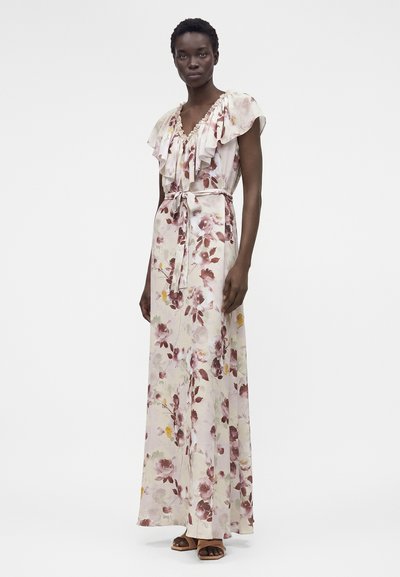 Γυναίκα που φοράει μακρύ, ανοιχτόχρωμο φόρεμα με floral μοτίβο, βολάν στο V-λαιμό και ζώνη στη μέση, στέκεται με ουδέτερη έκφραση και καφέ σανδάλια.