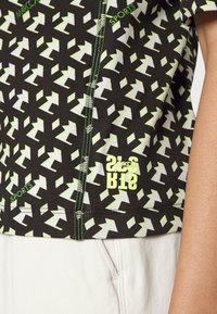 Top deportivo negro con estampado, que presenta diseños geométricos en verde y blanco, con un logo y detalles de costura en contraste. Fabricado en material acanalado.