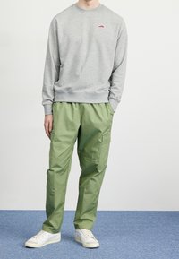 Grå sweatshirt med en liten röd logotyp, kombinerad med ljusgröna cargobyxor med fickor och vita sneakers på ett blått golv.