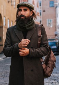 Manteau en laine marron avec boutons noirs, écharpe en laine verte et sac à bandoulière en cuir marron. Un homme se tient sur une rue en pavés, portant une casquette à motifs.