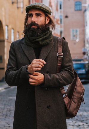Cappotto di lana marrone con bottoni neri, sciarpa lavorata a maglia verde e borsa a tracolla in pelle marrone. Un uomo si trova su una strada in ciottoli, indossando un berretto a fantasia.