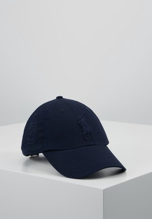 Casquette de baseball bleu foncé avec logo de joueur de polo brodé et numéro 3 sur le côté, posée sur une surface blanche devant un fond gris.
