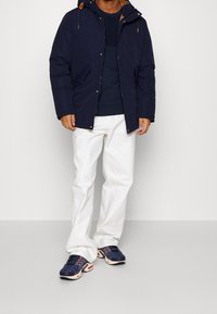 Marinblå pufferjacka med en kontrasterande brun foder, bärs över en marinblå stickad tröja, vita byxor och marinblå sneakers med orangea accenter.