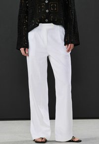 Schwarze offene Strickjacke mit einem verkürzten Design, ausgestattet mit großen Knöpfen, getragen über weißen, weit geschnittenen Hosen mit glatter Textur und maßgeschneiderter Passform.