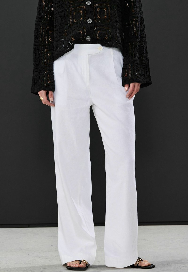 Schwarze offene Strickjacke mit einem verkürzten Design, ausgestattet mit großen Knöpfen, getragen über weißen, weit geschnittenen Hosen mit glatter Textur und maßgeschneiderter Passform.