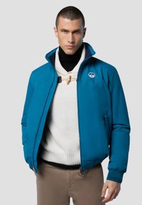 Giacca imbottita teal con colletto alto, chiusura a zip e patch con logo. Indossata sopra un maglione bianco a trama. I pantaloni marroni completano l'outfit.