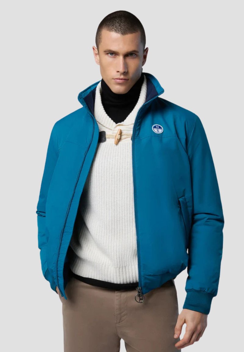 Giacca imbottita teal con colletto alto, chiusura a zip e patch con logo. Indossata sopra un maglione bianco a trama. I pantaloni marroni completano l'outfit.