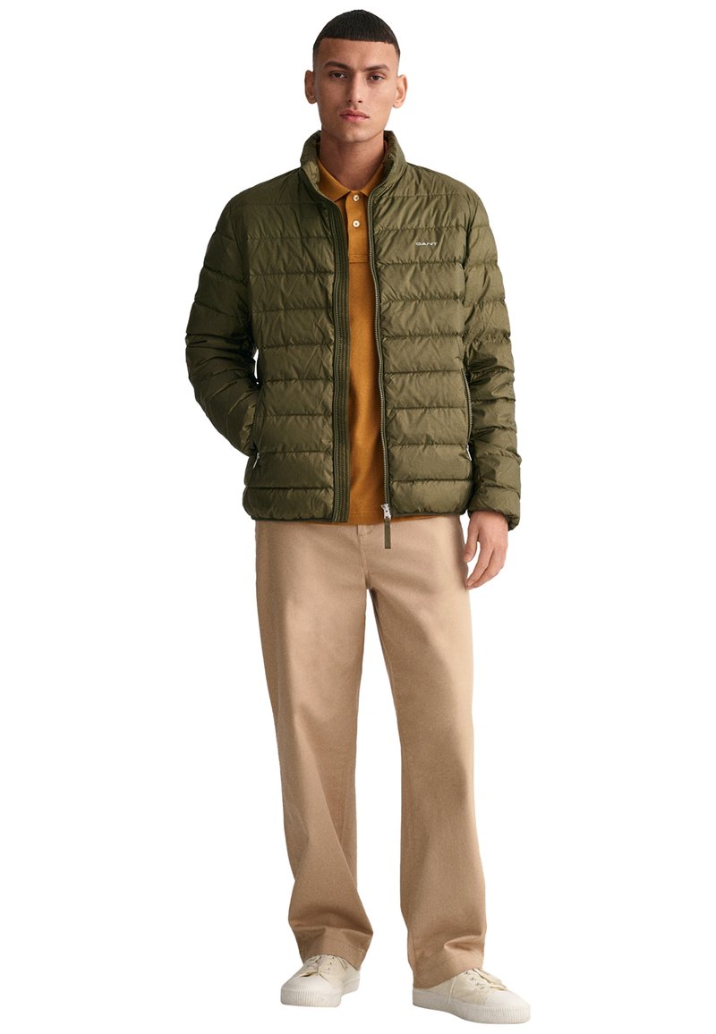 Chaleco de puffer verde con diseño de acolchado horizontal, cierre de cremallera y cuello alzado. Lleva puesto un polo marrón, combinado con pantalones beige y zapatillas blancas.