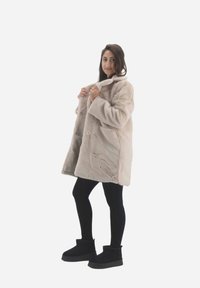 Beige faux fur coat med høj krave, knaplukning og sidelommer, båret over sorte leggings og sorte ankelsko. Glat tekstur.