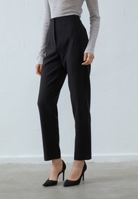 Pantaloni neri a vita alta con tasche laterali, fit sartoriale e pieghe pressate. Abbinati a décolleté nere a punta e a un top grigio chiaro.