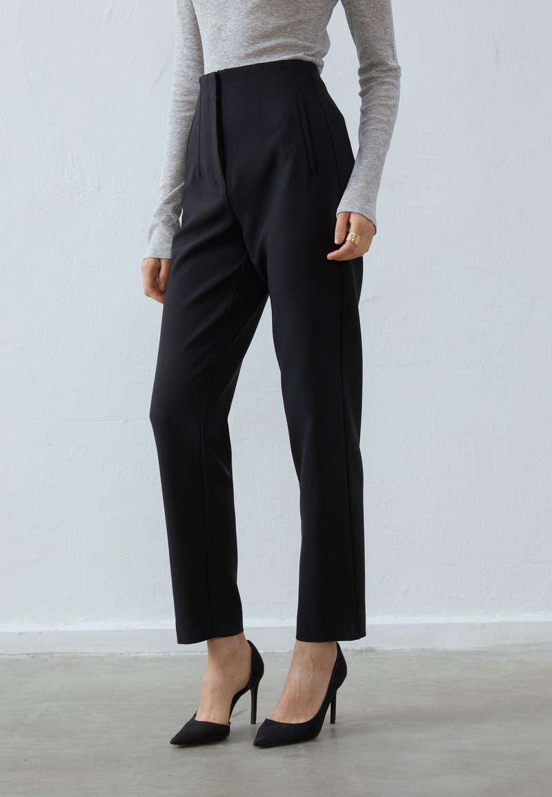 Pantaloni neri a vita alta con tasche laterali, fit sartoriale e pieghe pressate. Abbinati a décolleté nere a punta e a un top grigio chiaro.