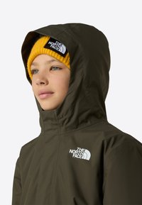 Geacă impermeabilă verde măsliniu cu glugă, având un logo alb. Combinată cu o căciulă tricotată galben intens, cu o etichetă.