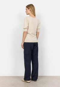 Pullover beige avec des manches bouffantes courtes, associé à un pantalon cargo bleu marine avec des poches latérales, et des chaussures plates de couleur claire.
