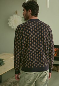 Marineblauer Pullover mit einem wiederholten goldenen Baum-Muster, gerippten Bündchen und Saum, kombiniert mit hellgrünen Jeans. Strukturierter Stoff.