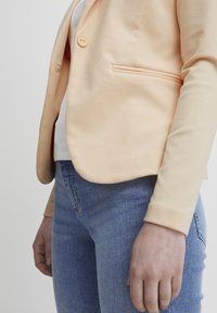 Béžový blazer s jedním knoflíkem, hladkým povrchem a boční kapsou. Nesený přes bílou košili, v kombinaci s modrými džínami.