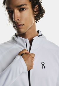 White zip-up jacket with a high collar, featuring a black logo on the chest and contrasting black zipper. Textured fabric detail on sleeves.