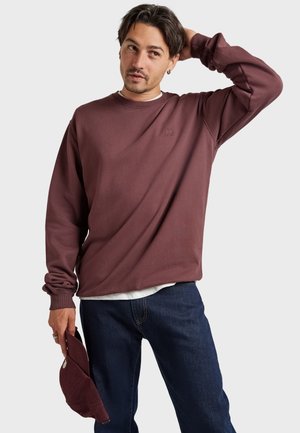 Homme portant un sweat-shirt marron à manches longues et un jean bleu, tenant une casquette marron assortie dans une main et touchant sa tête de l'autre.