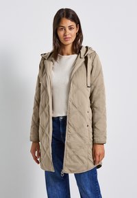 Beige gewatteerde lange jas met een capuchon, voorzien van drukknoopsluitingen, zijzakken en een gestructureerde matte afwerking. Gestyled met blauwe jeans.