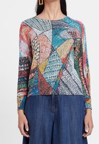 Camiseta de mangas largas multicolor con un diseño de patchwork que presenta diversas texturas y patrones, incluyendo formas geométricas y acentos metálicos.