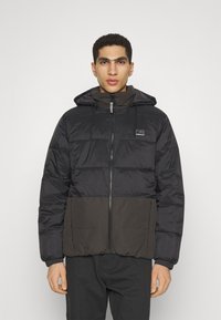 Black and dark gray puffer jacket with a hood, front zip closure, elastic cuffs, and logo detail on the left chest pocket.