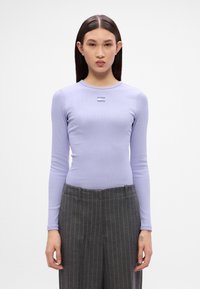 Langan ärmliches, helllila Oberteil mit runder Ausschnitt und geripptem Material. Es hat ein kleines Graphic auf der Brust. Kombiniert mit grauen Nadelstreifenhosen.