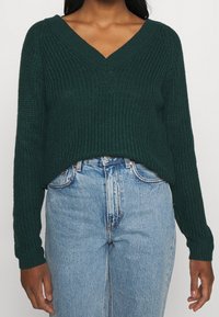 Dunkelgrüner gestrickter Pullover mit V-Ausschnitt und langen Ärmeln, der ein strukturiertes Muster und eine lässige Passform aufweist, kombiniert mit hellblauen Jeans.