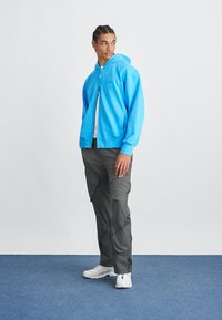 Ανοιχτό μπλε φούτερ με φερμουάρ και κουκούλα, συνδυασμένο με σκούρες γκρι cargo παντελόνες με τσέπες και ρυθμιζόμενους ιμάντες, φορεμένο με λευκά αθλητικά παπούτσια.