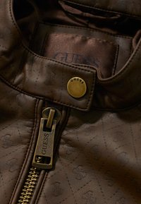 Gros plan sur une veste en cuir marron avec une fermeture éclair en laiton et un bouton-pression, tous deux marqués "GUESS", présentant un motif subtilement embossé sur le cuir.