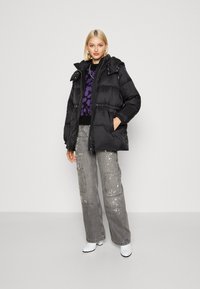Blouson noir avec capuche, présentant une taille cintrée et une fermeture éclair. Associé à un pantalon gris avec des éclaboussures de peinture blanche et des chaussures blanches.