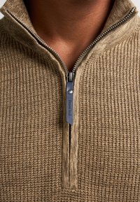 Ruskea neulepusero, jossa on neljännesvetoketju. Teksturoitu kangas, ribattu kaulus ja tumma vetoketjun vetopidike, jossa on logo. Lähiökuva.