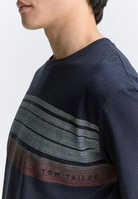 Marineblaues T-Shirt mit kurzen Ärmeln, das horizontale Streifenmuster in verschiedenen Farbtönen zeigt und den Text "TOM TAILOR" nahe dem Saum trägt. Weicher Stoffeindruck.