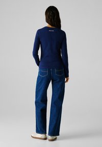 Maglietta a maniche lunghe blu mare in morbido tessuto, con scollatura rotonda e logo "PEPE JEANS" sul retro. Abbinata a jeans blu a vestibilità ampia.