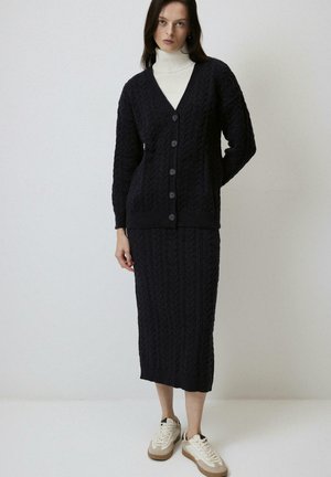 Navy cable-knit cardigan with buttons, paired with a matching cable-knit midi skirt. The model wears white turtleneck and beige sneakers.