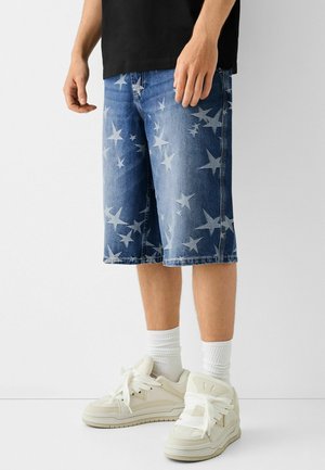 Man met blauwe denim shorts met witte sterpatronen, witte crew-sokken, witte sneakers en een zwart T-shirt, staande op een witte vloer.