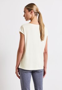 Top corto de color amarillo claro con cuello redondo, textura suave y dobladillo recto. Combinado con vaqueros grises, visto desde atrás.