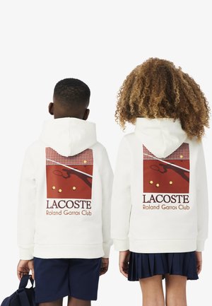 Due bambini di spalle indossano felpe con cappuccio bianche Lacoste Roland Garros Club con una grafica di campo da tennis e giocatore sul retro.