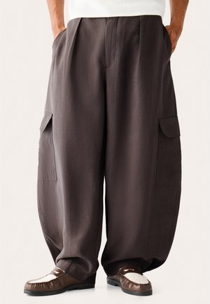 Man draagt losse donkerbruine cargobroek met zijzakken en bruin-witte loafers, handen in de zakken.