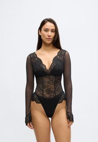 Črn čipkasti bodi z prosojnimi dolgimi rokavi, z globokim V-izrezom in cvetličnimi vzorci. Oblikovanje dopolnjuje ščetinasto rob.