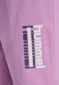 Close-up of purple sportswear fabric with "hummel SPORTSWEAR" logo in white and pink vertical text on black bars.