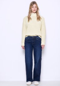 Maglione a collo alto a coste crema abbinato a jeans a gamba larga blu scuro con strisce laterali bianche e scarpe neutre. Design semplice e moderno.