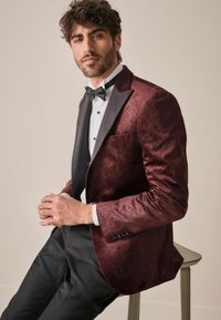 Sacou din catifea bordeaux cu un model texturat în diamante, rever din satin negru, cămașă albă și papion negru. Pantaloni slim-fit completează ținuta.
