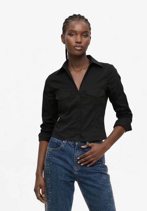 Kvinde iført en tætsiddende sort skjorte med knapper og blå jeans med rhinsten detaljer, stående med den ene hånd i lommen mod hvid baggrund.