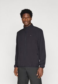 Giacca nera con zip, colletto alto, polsini elasticizzati, due tasche laterali e un piccolo logo CK sul petto. Tessuto liscio e leggero.