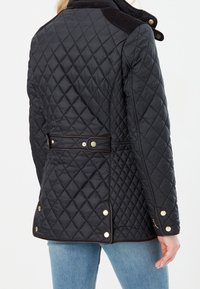 Woman wearing black quilted jacket with gold buttons and adjustable back belt, paired with light blue jeans, shown from behind.