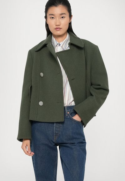 Green cropped wool coat with a wide collar, double buttons, and side pockets, paired with a striped shirt and dark denim jeans.