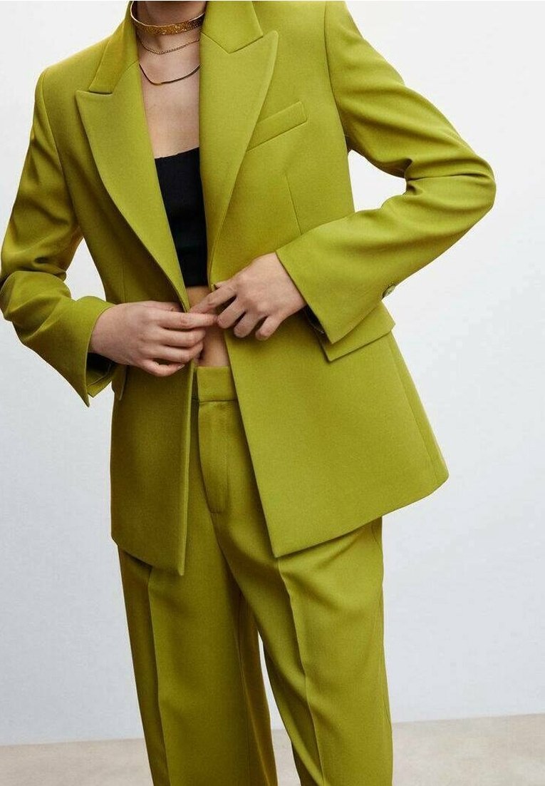 Personne boutonnant une veste de costume vert citron sur mesure par-dessus un pantalon assorti, portant un top court noir et des colliers en or superposés.